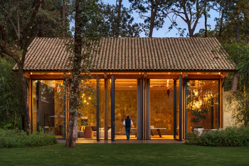 Casa entre árboles al atardecer con grandes puertas vidriadas iluminadas